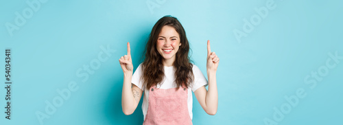 Image of happy smiling young woman with curly hair, laughing and pointing fingers up at logo banner, showing promo deal, standing against blue background