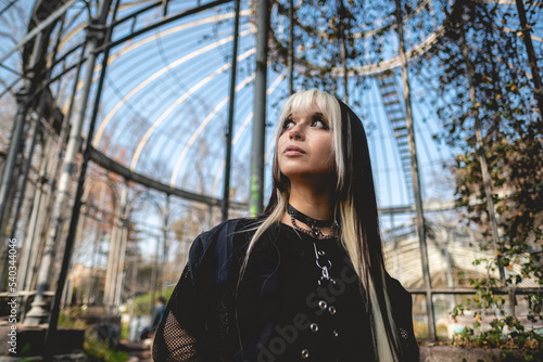 Young and skinny model with long blonde and black hair and black clothes and makeup posing inside an old abandoned greenhouse