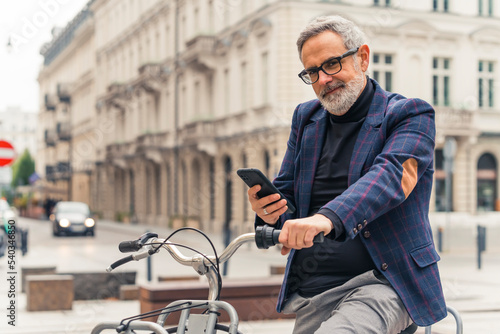 Modern bearded grey-haired mature man using his smartphone and riding a bicycle in the city streets with beautiful buildings in the background. High quality photo