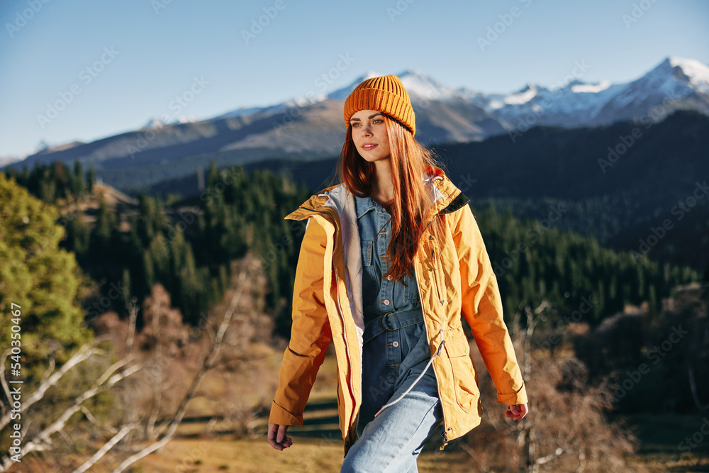 Woman hiking in the mountains in the fall with a smile with teeth and happiness in a yellow cape with red hair full-length stands against the backdrop of trees and mountains in the sunset light