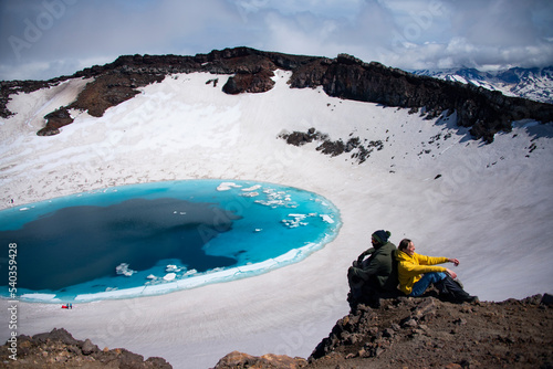 A lake in a crater
Man and woman are sitting on the edge of crater of Gorely volcano. There’s a light blue lake in the background and snow on walls of the crater. photo