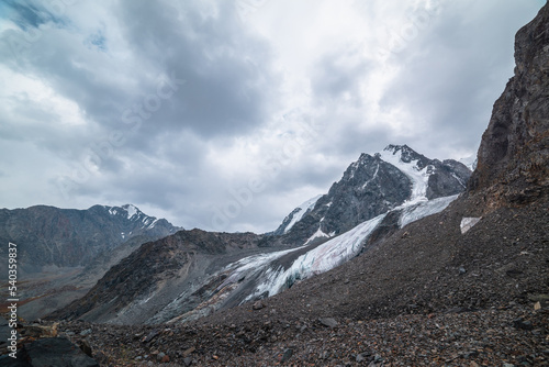 Dramatic landscape with hanging glacier against large snow mountain with sharp rocky pinnacle under gray cloudy sky. Glacier with icy cornice in high altitude. Gloomy scenery in mountains in overcast.