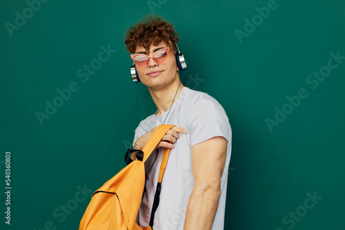 a funny man stands on a gray background in pink glasses and looks into the camera listening to music, holding an orange backpack in his hand
