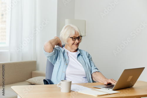 a happy, relaxed elderly woman works from home at a laptop and happily looking at the monitor holds her hand behind her head