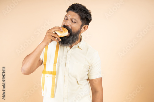 South indian man wearing traditional white dress eating burgers fast food or junk food snack isolated on beige background
