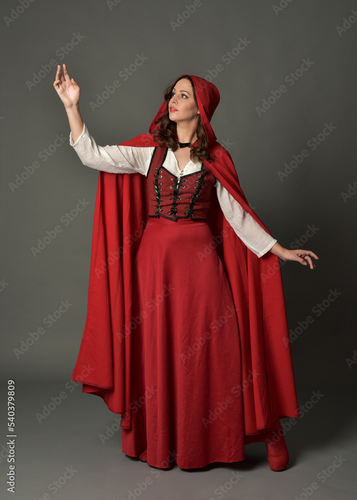 Obraz premium Full length portrait of beautiful brunette woman wearing red medieval fantasy costume with long skirt and flowing hooded cloak. Standing pose with gestural hand poses, isolated on grey studio backgrou