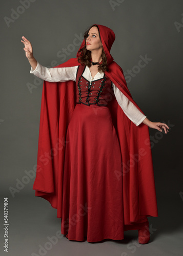 Full length portrait of beautiful brunette woman wearing red medieval fantasy costume with long skirt and flowing hooded cloak. Standing pose with gestural hand poses, isolated on grey studio backgrou