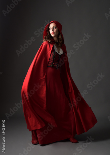 Full length portrait of beautiful brunette woman wearing red medieval fantasy costume with long skirt and flowing hooded cloak. Standing pose with gestural hand poses, isolated on grey studio backgrou