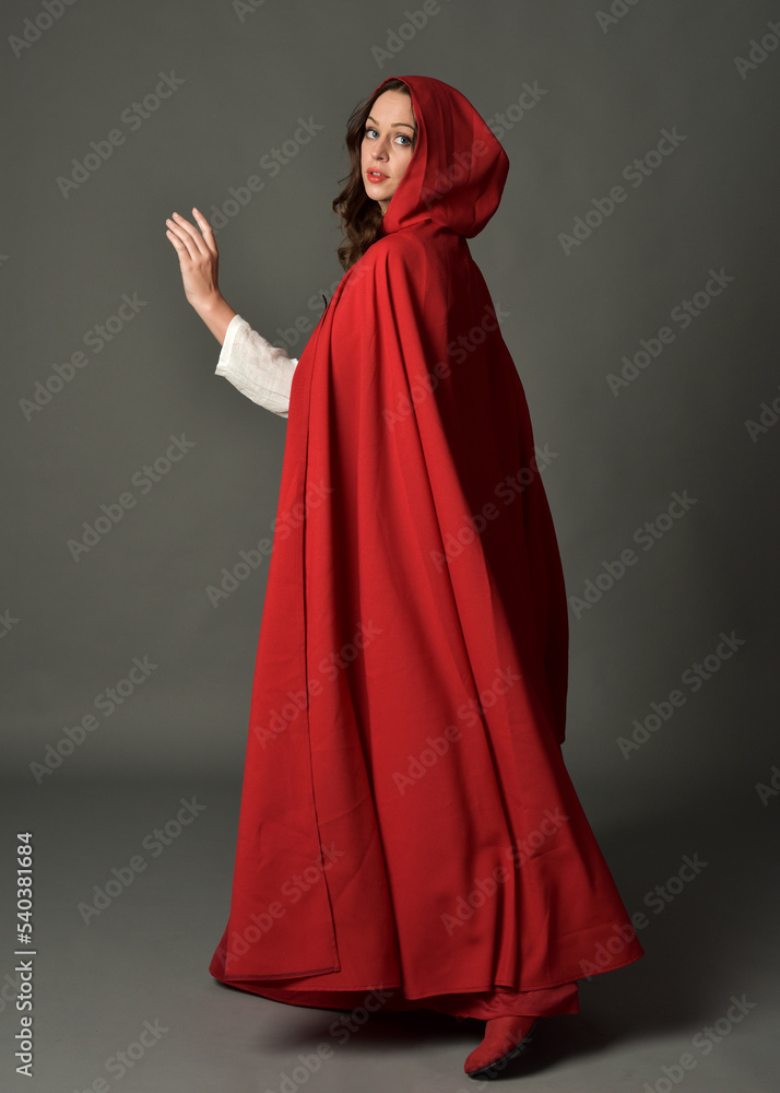 Full length portrait of woman wearing red medieval fantasy costume, flowing hooded cloak. Standing pose in backview, gestural hand poses, walking away from camera isolated on grey studio background. Full length portrait of woman wearing red medieval fantasy costume, flowing hooded cloak. Standing pose in backview, gestural hand poses, walking away from camera isolated on grey studio background.