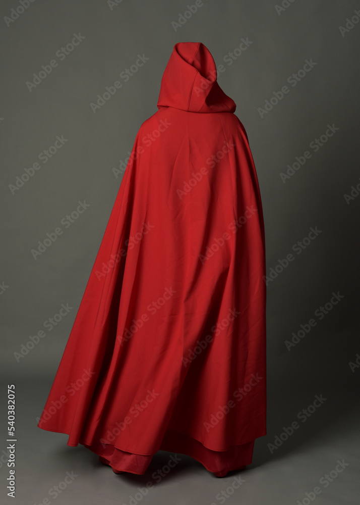 Full length portrait of woman wearing red medieval fantasy costume, flowing hooded cloak. Standing pose in backview, gestural hand poses, walking away from camera isolated on grey studio background.