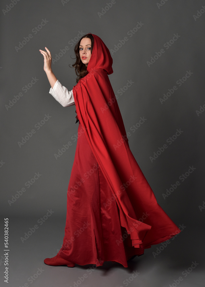 Full length portrait of woman wearing red medieval fantasy costume, flowing hooded cloak. Standing pose in backview, gestural hand poses, walking away from camera isolated on grey studio background.