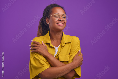Young happy beautiful African American woman millennial with curly hair hugs herself to cheer up and get rid of loneliness dressed in casual yellow shirt, stands on isolated purple background.