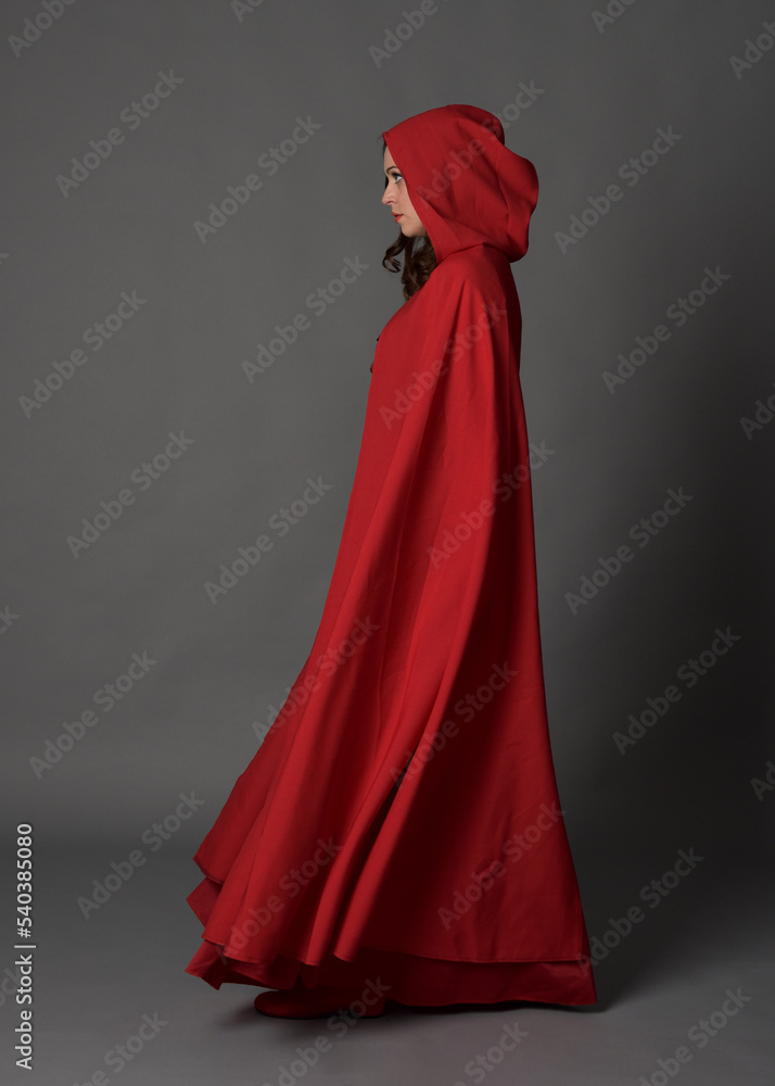 Full length portrait of woman wearing red medieval fantasy costume, flowing hooded cloak. Standing pose side profile, gestural hand poses, walking away from camera isolated on grey studio background. Full length portrait of woman wearing red medieval fantasy costume, flowing hooded cloak. Standing pose side profile, gestural hand poses, walking away from camera isolated on grey studio background.
