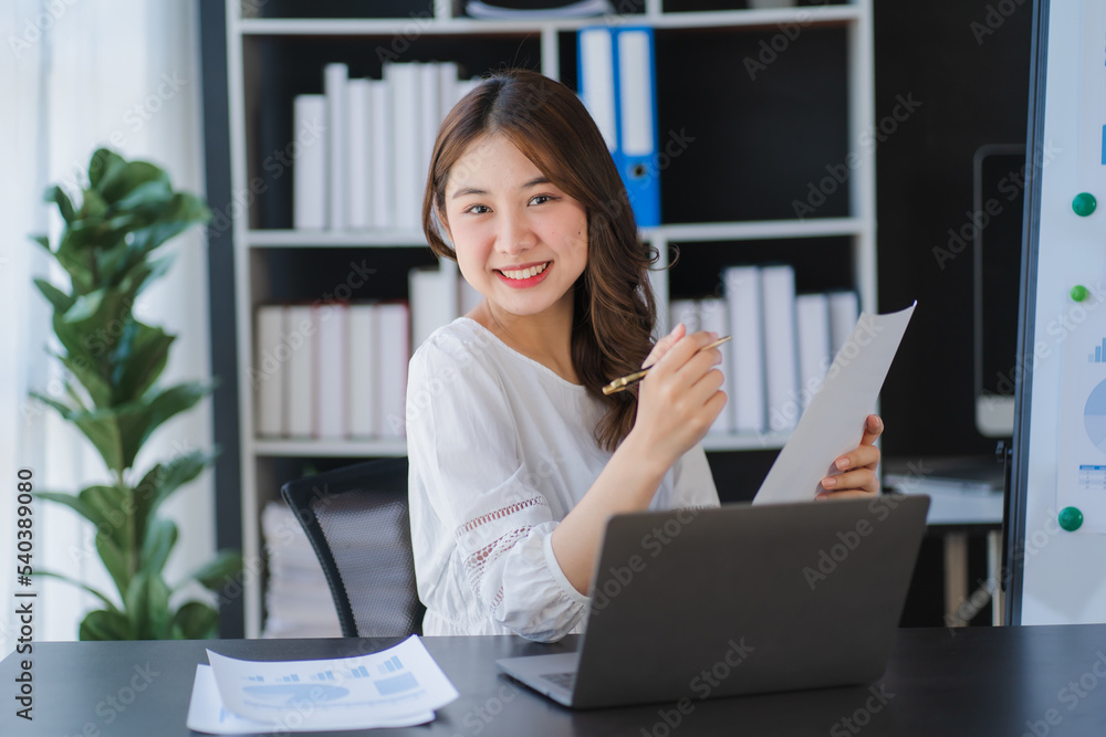 Beautiful Asian businesswoman working on a desk with a laptop with a smiling face while looking at the document received in the office. Beautiful Asian businesswoman working on a desk with a laptop with a smiling face while looking at the document received in the office.