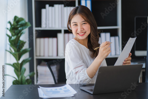 Beautiful Asian businesswoman working on a desk with a laptop with a smiling face while looking at the document received in the office.