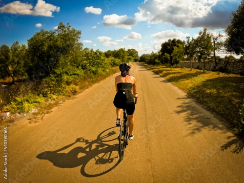 Back shot of an adult woman riding a bicycle on a lane through olive groves in Puglia, Italy