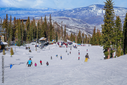 View of ski slope in Colorado, USA, in the winter; skiers and snowboarders skiing down to  chairlift; mountain range in background photo