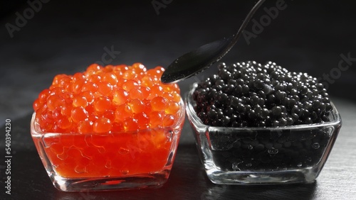 Red and black caviar in two identical transparent glass bowls on a black background. Man spoons caviar