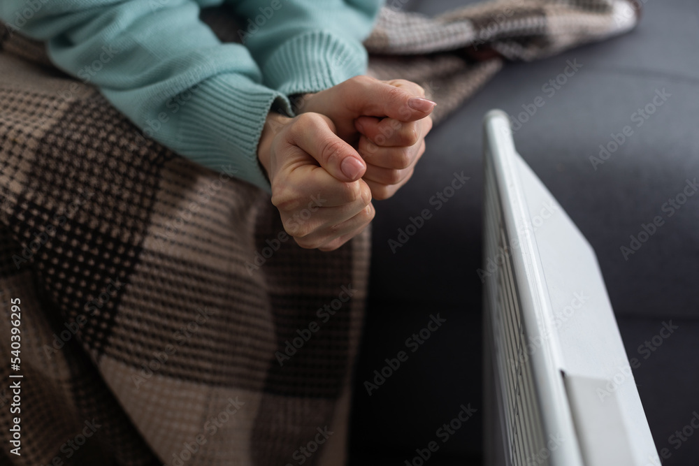 Girl warms up the frozen hands above hot radiator, close up. Woman wearing woolen sweater warming up while sitting near a heating radiator. Woman warming hands near electric heater at home.