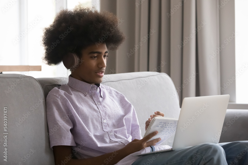 Attentive African student guy in headphones, holds copybook take notes, make assignment with classmate use video call, listen on-line tutor studying sit on sofa at home with laptop on laps. Education Attentive African student guy in headphones, holds copybook take notes, make assignment with classmate use video call, listen on-line tutor studying sit on sofa at home with laptop on laps. Education