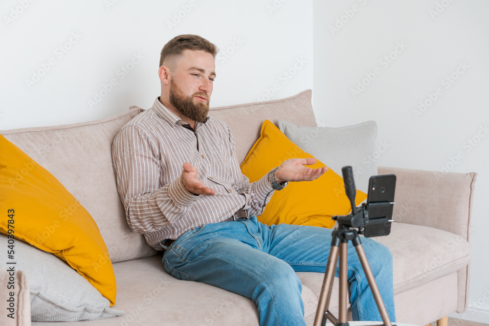 Smiling bearded guy filming video using smartphone on tripod online while sitting on sofa in living room wearing shirt and jeans. Social media influence, content creation and online consultation
