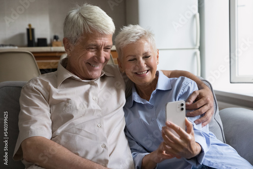 Attractive senior couple looking at cellphone screen while calling to grown up children via videocall mobile application, choose goods, buy services use electronic commerce web app. Modern tech usage