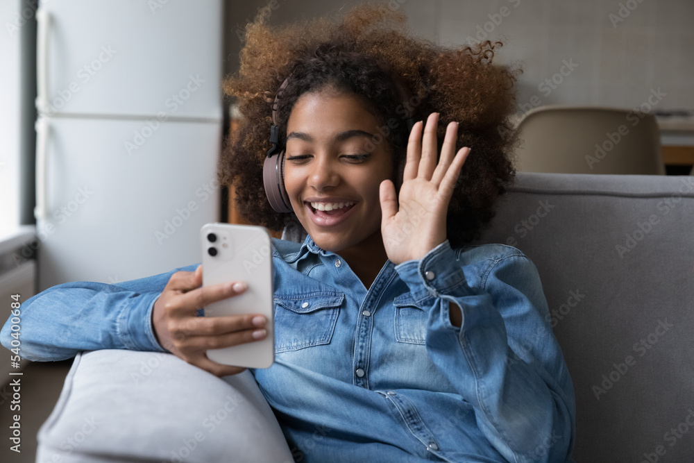 Φωτογραφία Attractive African teenager girl in wireless headphones greets friend start video call talk using smartphone seated on sofa at home