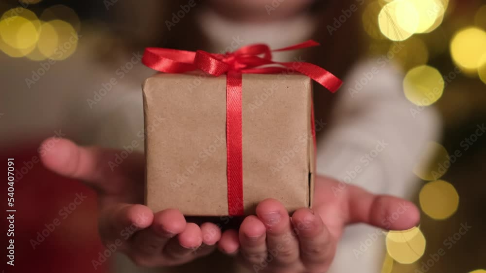Christmas gift close up. Kid hands holding small present box with red ribbon on background of sparkling lights. Child giving gift. Holidays and celebration concept Christmas gift close up. Kid hands holding small present box with red ribbon on background of sparkling lights. Child giving gift. Holidays and celebration concept
