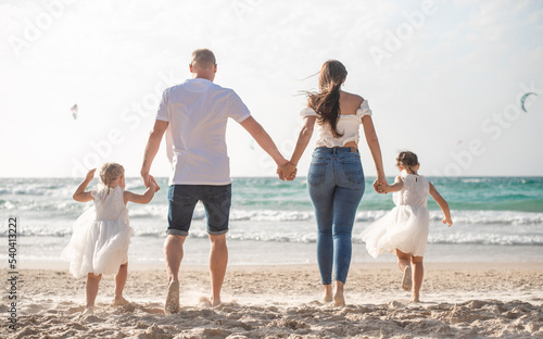 Happy family, back view. parents and two daughters on beach in summer vacation. Run, play, smile and get fun on sand near sea together. white clothes and jeans.