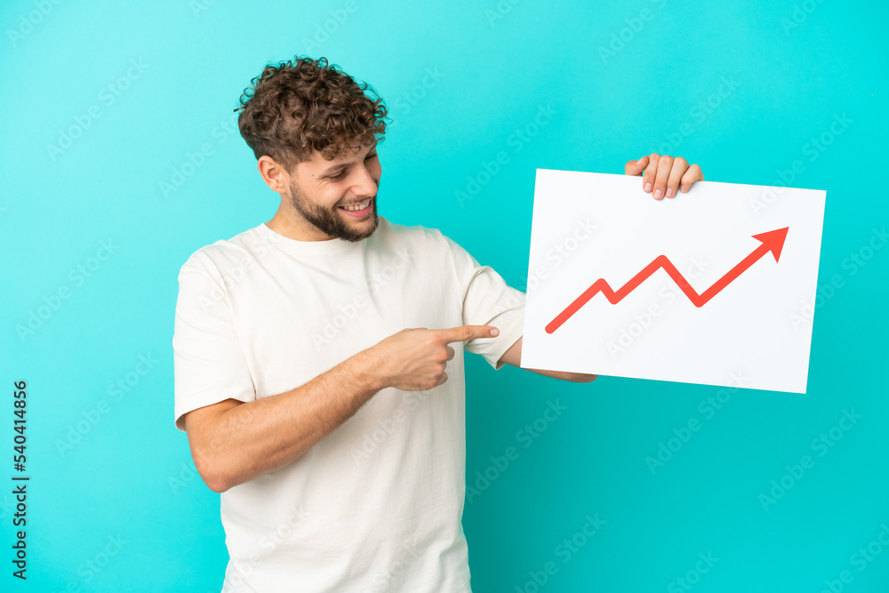 Young handsome caucasian man isolated on blue background holding a sign with a growing statistics arrow symbol and pointing it Young handsome caucasian man isolated on blue background holding a sign with a growing statistics arrow symbol and pointing it