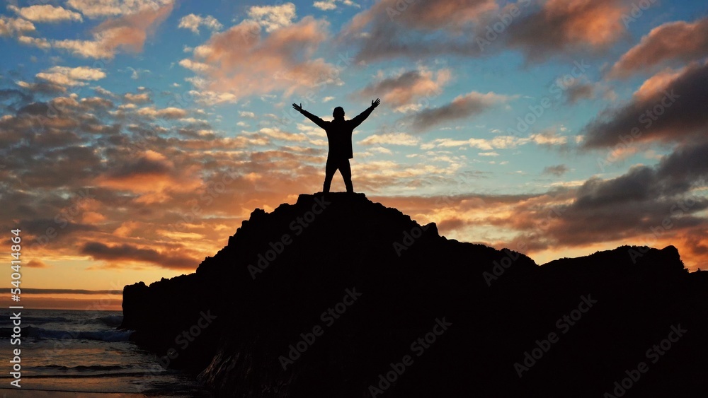 Man stands on a rock against the ocean during summer sunset. Human with raised hands looks to sun over horizon in morning while sunrise. Man feels happy and freedom. Silhouette of a man against  sky