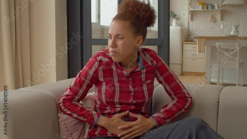 Portrait of unwell displeased beautiful black woman sitting on sofa, feeling discomfort in stomach and suffering from gynecological pain pressing crotch lower abdomen indoors.