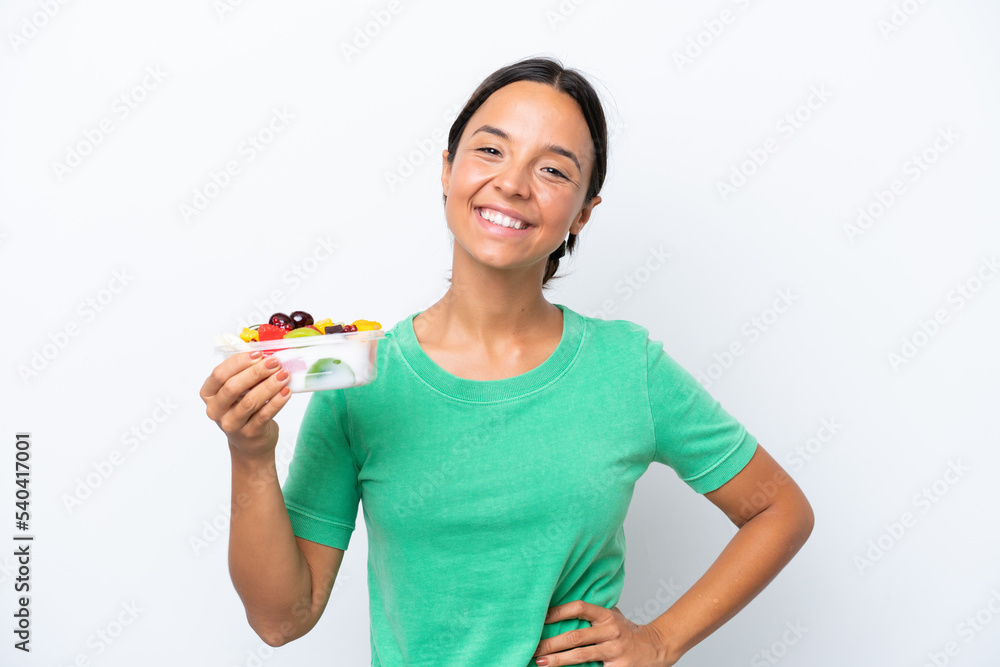 Young hispanic woman holding a bowl of fruit isolated on white background posing with arms at hip and smiling Young hispanic woman holding a bowl of fruit isolated on white background posing with arms at hip and smiling