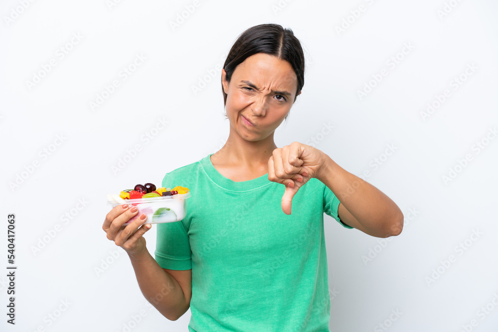 Young hispanic woman holding a bowl of fruit isolated on white background showing thumb down with negative expression Young hispanic woman holding a bowl of fruit isolated on white background showing thumb down with negative expression