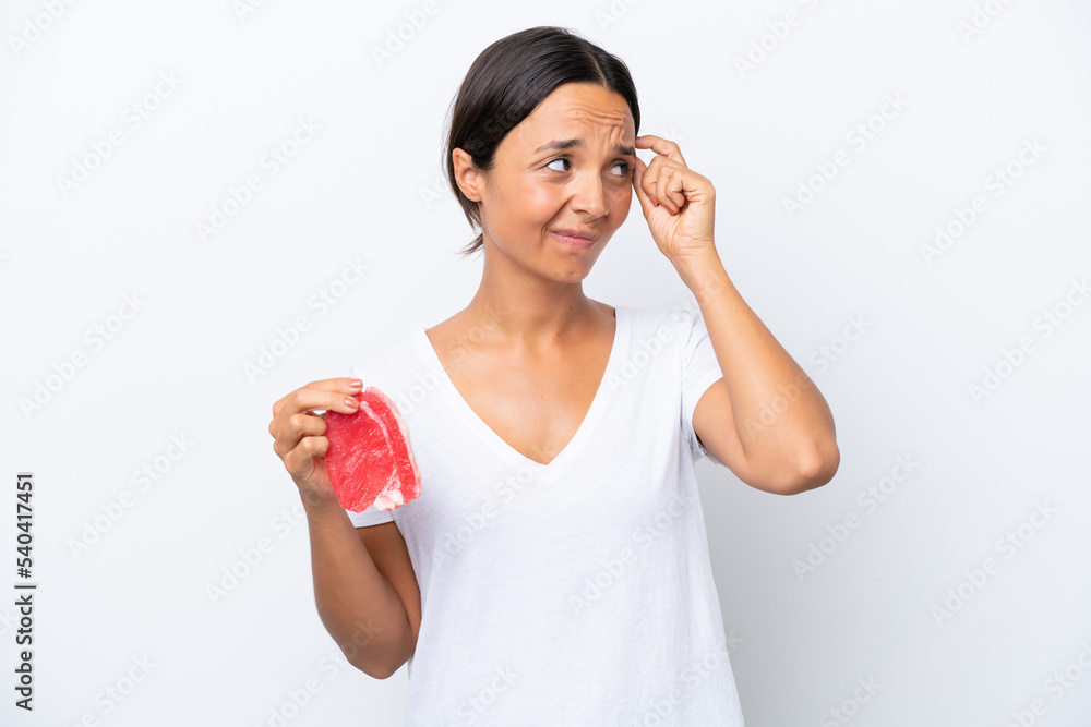 Young hispanic woman holding a piece of meat isolated on white background having doubts and with confuse face expression Young hispanic woman holding a piece of meat isolated on white background having doubts and with confuse face expression