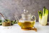 Fennel tea in a glass teapot, fresh fennel bulb, fennel seeds on white wooden table with grey background