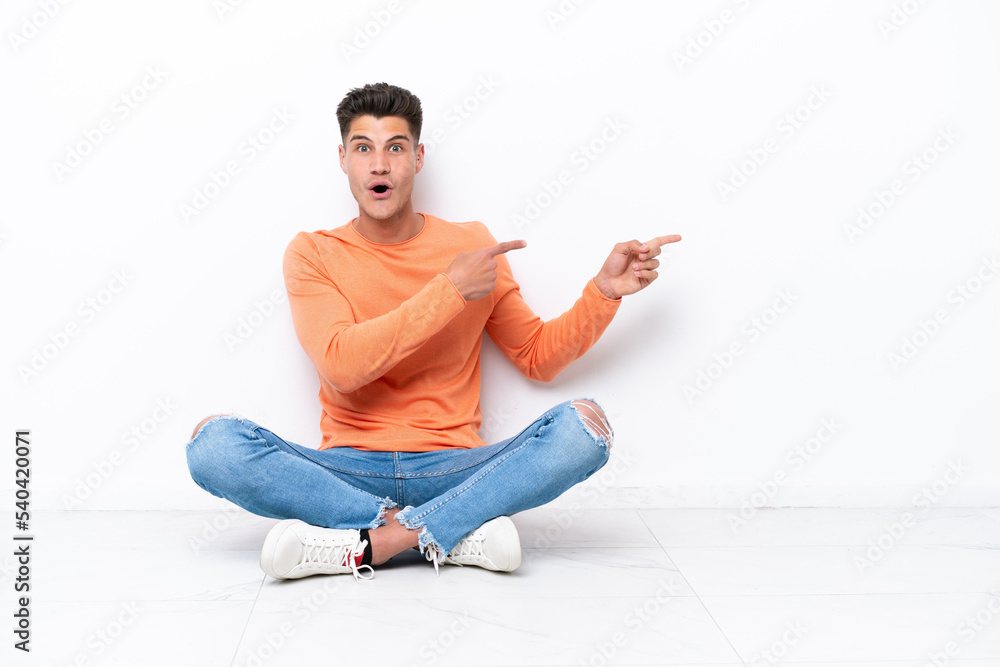 Young man sitting on the floor isolated on white background surprised and pointing side