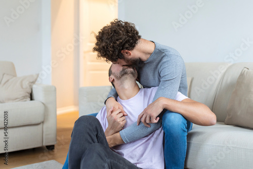 Lovely gay couple kissing while sitting in their living room at home. Two romantic young male lovers having fun indoors. Young gay couple living together.