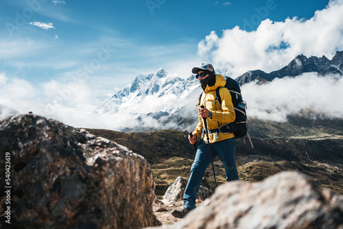 Outdoor tourist traveling along high altitude mountains wearing yellow jacket and professional backpack. Young solo hiker walk across sunny mountain track