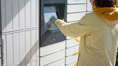 Young woman choosing operation touch screen of outdoor automated parcel machine or locker to deposit the parcel for storage, wearing yellow clothes. Back view. Mail delivery and post service concept.