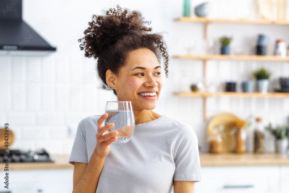 Healthy lifestyle concept. Beautiful young African American woman holding glass of fresh clean water standing in the home kitchen, looking at away and smiles friendly