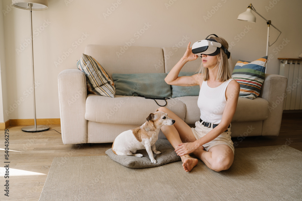 Virtual reality at home. Blonde woman testing VR glasses headset watching  360 degree video. Long slim naked legs. Small dog Jack Russell terrier. Be  present in the moment escaping from reality foto