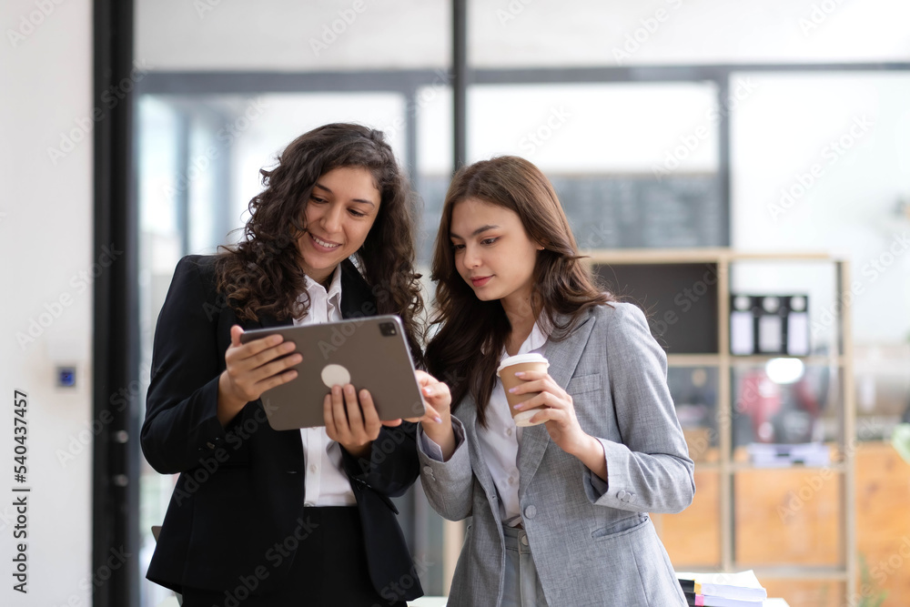 Obraz premium Two beautiful Asian businesswomen standing using digital tablet consulting and analyzing information in office work.