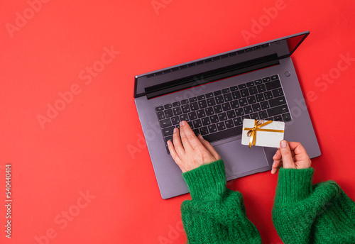 Christmas shopping concept. Flat lie. Women's hands in a green sweater on laptop keyboard with Gift card on a red background photo