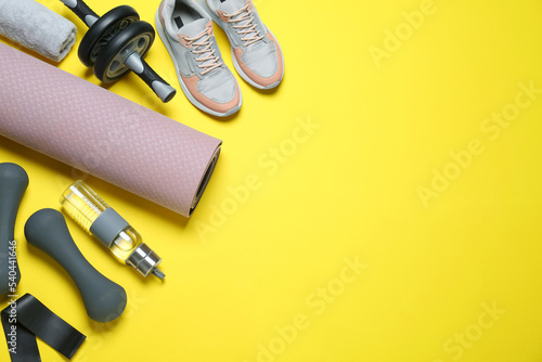 Exercise mat, dumbbells, bottle of water, ab roller, fitness elastic band, towel and shoes on yellow background, flat lay. Space for text