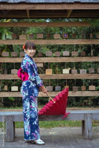 Japanese women with beautiful dresses casual Yukata Kimono and red umbrellas wear traditional clothes and stand with hold a red umbrella and wall for hanging wooden signs wishing sacred things.