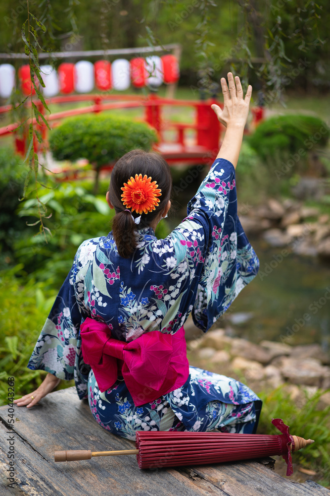 Japanese women with beautiful dresses casual Yukata Kimono and red umbrellas wear traditional clothes and sit on a wooden chair relaxing in the atmosphere fresh natural garden. Japanese women with beautiful dresses casual Yukata Kimono and red umbrellas wear traditional clothes and sit on a wooden chair relaxing in the atmosphere fresh natural garden.
