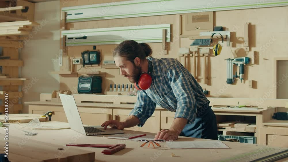 Attractive Carpenter Using a Laptop Making an Online Project in a Loft Studio with Tools on the Walls. Woodworking. Manual Labor. Factory. Carpentry, Craftsmanship, and Handwork Concept. Attractive Carpenter Using a Laptop Making an Online Project in a Loft Studio with Tools on the Walls. Woodworking. Manual Labor. Factory. Carpentry, Craftsmanship, and Handwork Concept.