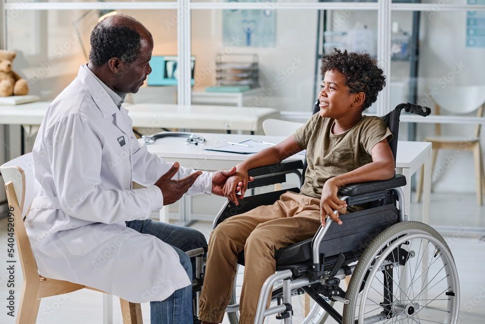 African doctor in white coat talking to little boy with disability during his visit at clinic