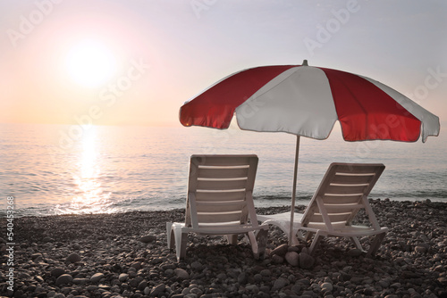 Fototapeta Naklejka Na Ścianę i Meble -  Empty sunbeds and umbrella on stone beach near sea at sunset